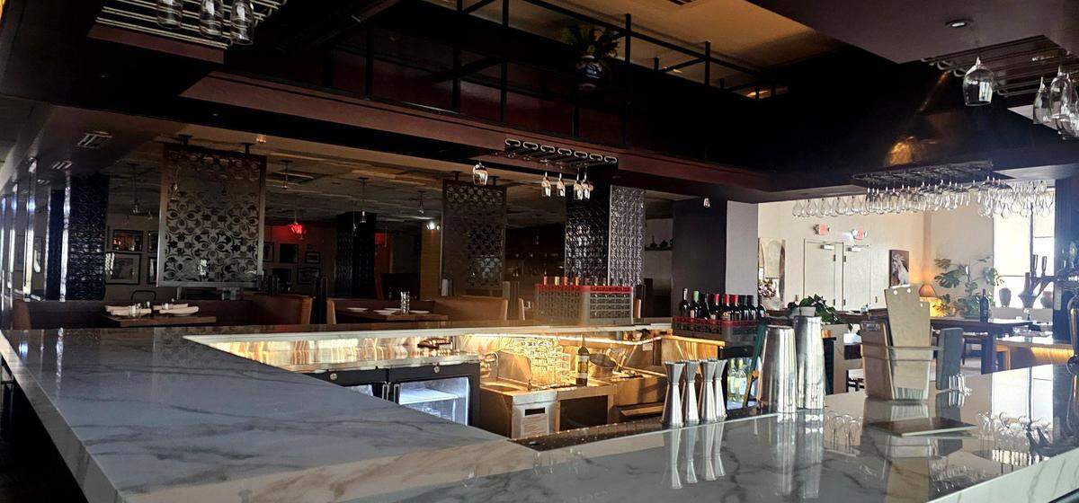 A wide, eye-level shot of a restaurant’s bar area. The long counter features a white marble top, and the service area holds cocktail shakers and bottles. Mirrored decorative screens and dark wood paneling separate the bar from the dimly lit dining room in the background.