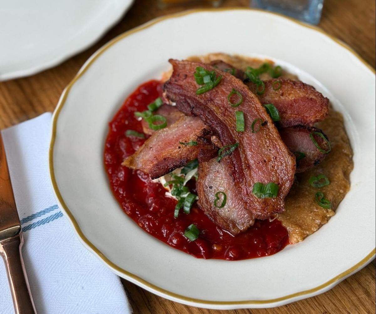 A close-up, high-angle shot of a dish served on a white plate with a gold rim. The plate holds brown grits and a pool of bright red sauce, topped with three slices of thick-cut crispy bacon and chopped green onions.