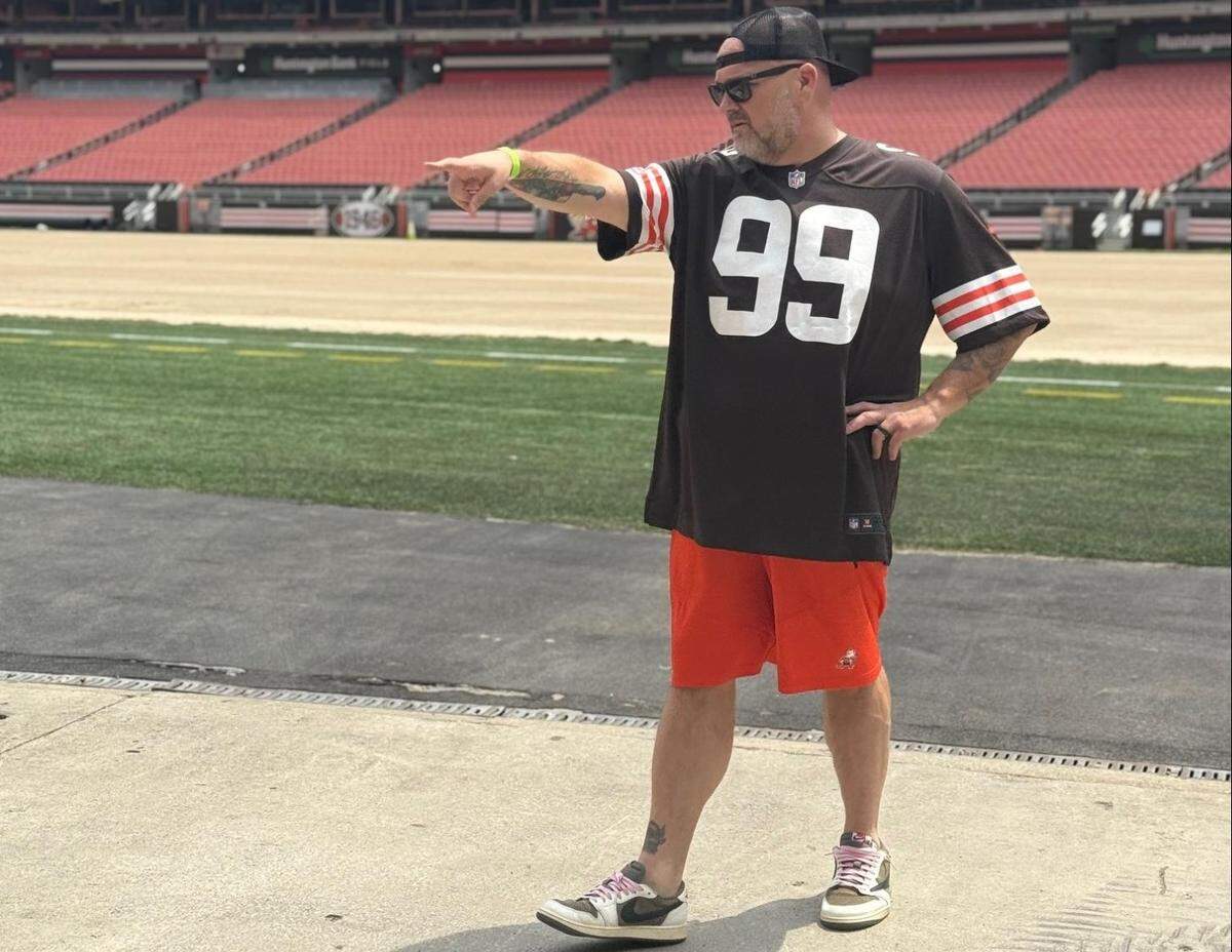A man with a beard, a black baseball cap, and sunglasses is standing on the sidelines of a football field. He is wearing a black jersey with the number “99” in white, red shorts, and sneakers. With one hand on his hip, he points his other arm and finger out to the left, looking in the same direction. In the background, the empty red stadium seating is visible under a sunny sky.