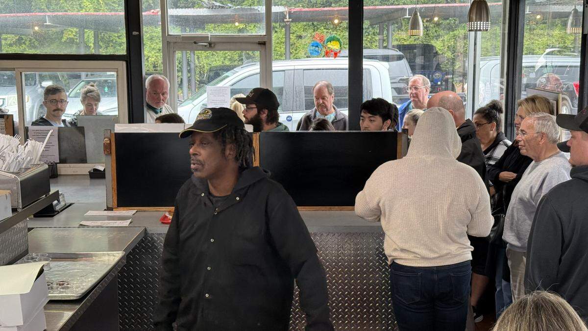 An interior view of a restaurant from behind a stainless-steel service counter. In the foreground, a staff member works at the counter. In the background, a long line of customers is visible, stretching along a wall of windows and out the glass door.