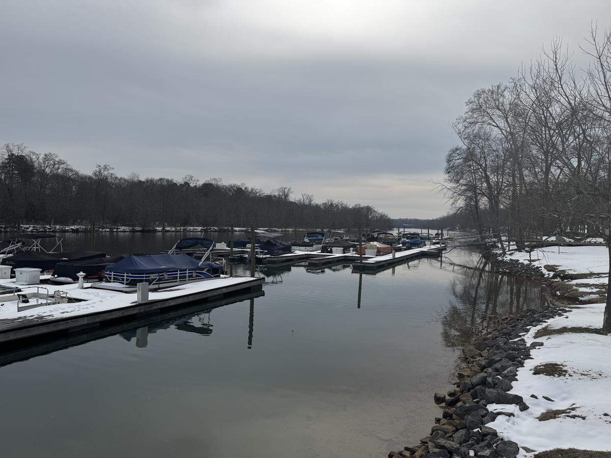 A quiet marina on a river during winter, with several pontoon boats docked and covered in blue tarps. The riverbanks and docks are covered in a thin layer of snow under an overcast sky.