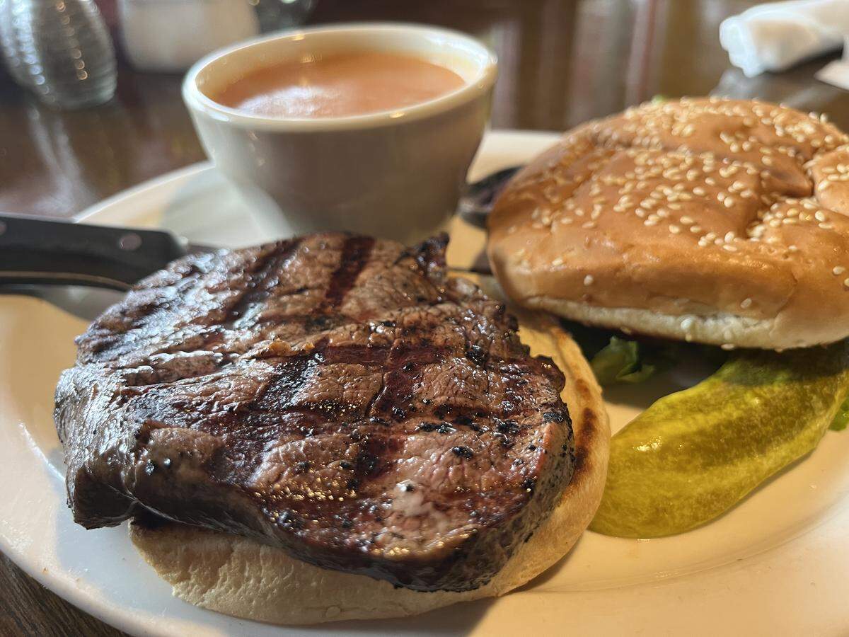 A close-up shot of a restaurant meal on a white plate. The main item is a steak sandwich, with a thick, round grilled steak showing dark char lines resting on the bottom half of a bun. The top half of the bun, covered in sesame seeds, is next to it. A large pickle spear and a white cup of orange-colored soup complete the meal.
