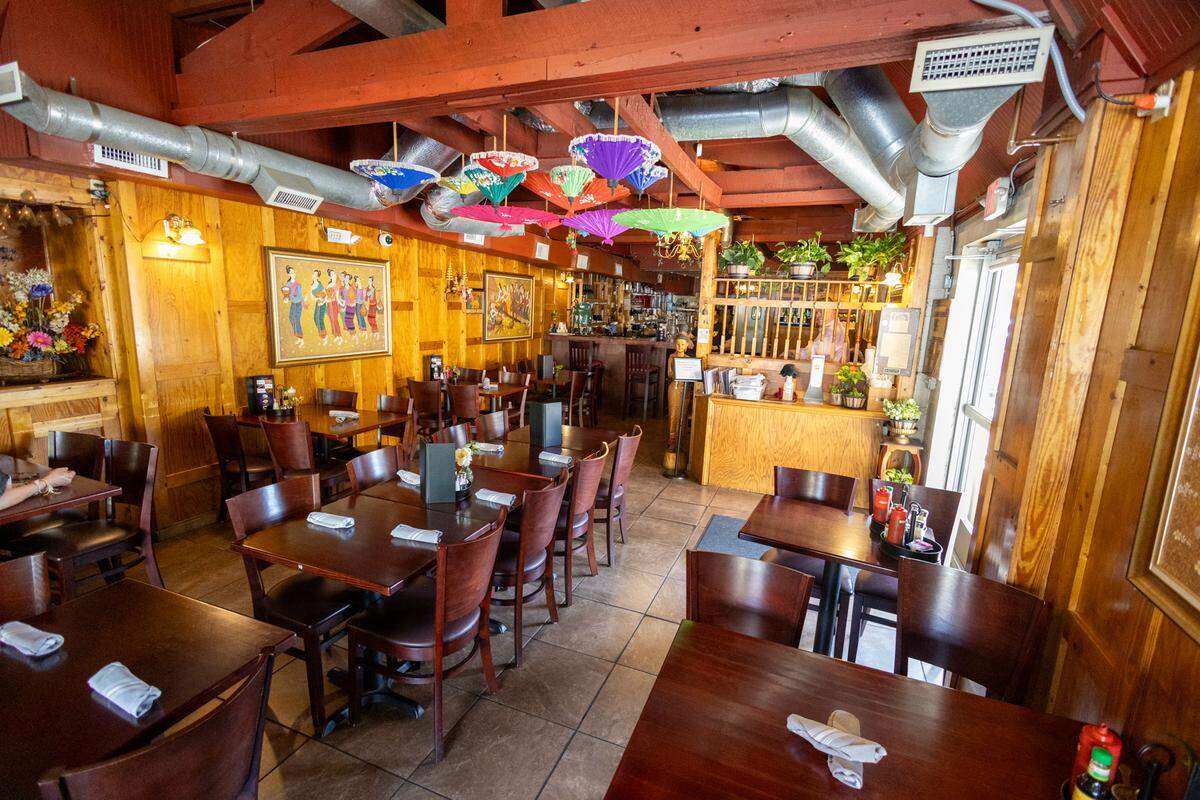 A wide view of a restaurant dining room featuring warm wood-paneled walls, dark wooden tables, and exposed ceiling beams. Colorful traditional Thai umbrellas and greenery hang from the ceiling, adding a vibrant touch to the rustic space.