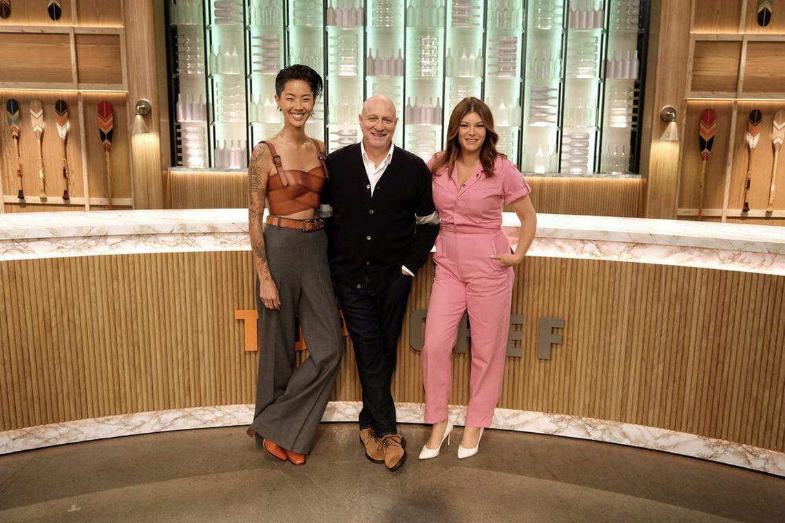 Three people, two women and one man, stand side-by-side in front of a modern, wood-paneled set that resembles a kitchen or studio. The woman on the left has short, dark hair and is smiling at the camera, wearing a brown leather-look bralette and loose grey pants with orange shoes. The man in the center is bald and smiling, dressed in a black cardigan over a white shirt and dark pants. The woman on the right has long brown hair and is smiling, wearing a pink jumpsuit and white heels, with her hands in her pockets. Behind them, there are shelves with decorative paddles or oars on the left and a wall of illuminated, vertically-oriented panels on the right. A curved counter with a wood slatted front is in the foreground.