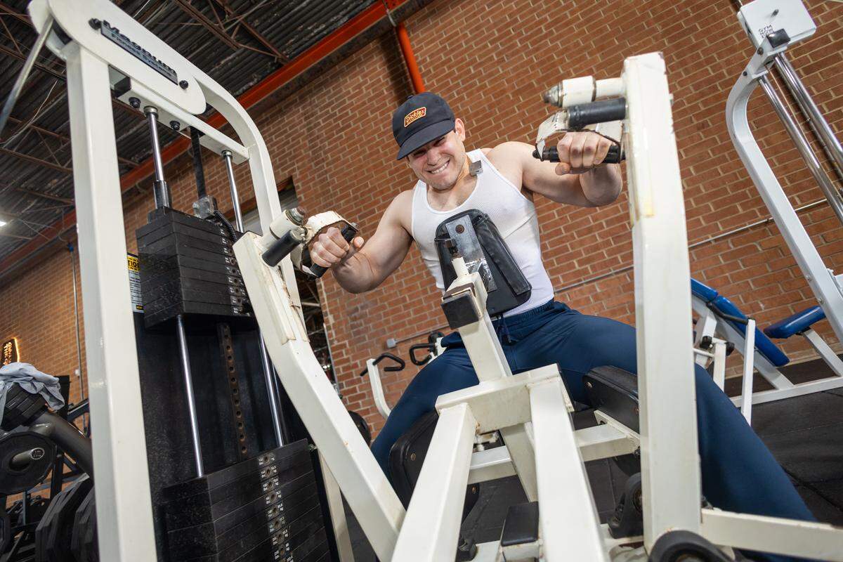 Close-up of a gym user in a white tank top performing a chest exercise on a white, vintage weight machine against a red brick wall background.