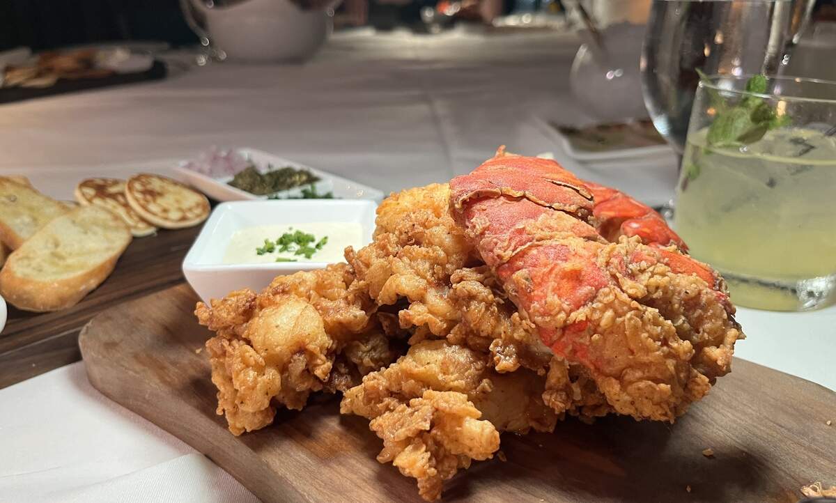A heap of crispy, golden fried lobster meat served on a wooden board alongside a bright red lobster tail shell, a small white bowl of dipping sauce topped with chives, sliced crostini, and a mint cocktail on a white tablecloth.