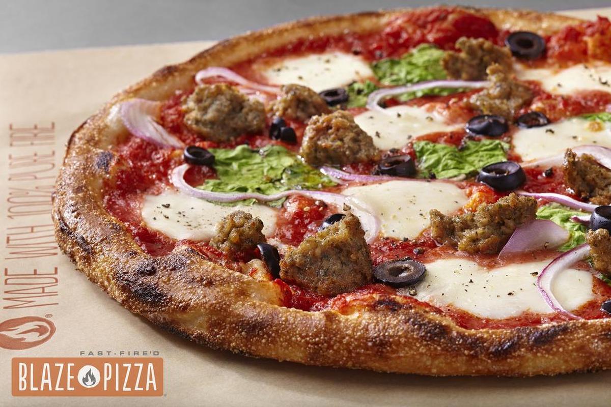 A detailed shot of a custom-built pizza, possibly from Blaze Pizza, highlights a well-charred crust and fresh toppings. The pizza features a red sauce base with fresh mozzarella, crumbles of sausage, black olive slices, and thin rings of red onion and fresh spinach leaves.