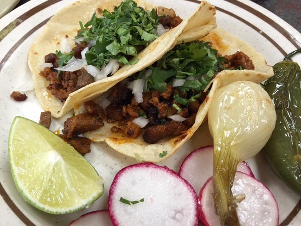 A close-up, overhead shot shows two corn tortillas, partially folded, filled with seasoned meat, diced white onions, and chopped fresh cilantro. They are served on a white plate with brown stripes around the rim. Accompanying the tacos are a wedge of lime, several slices of red radish, a roasted whole onion, and a roasted green jalapeño pepper.