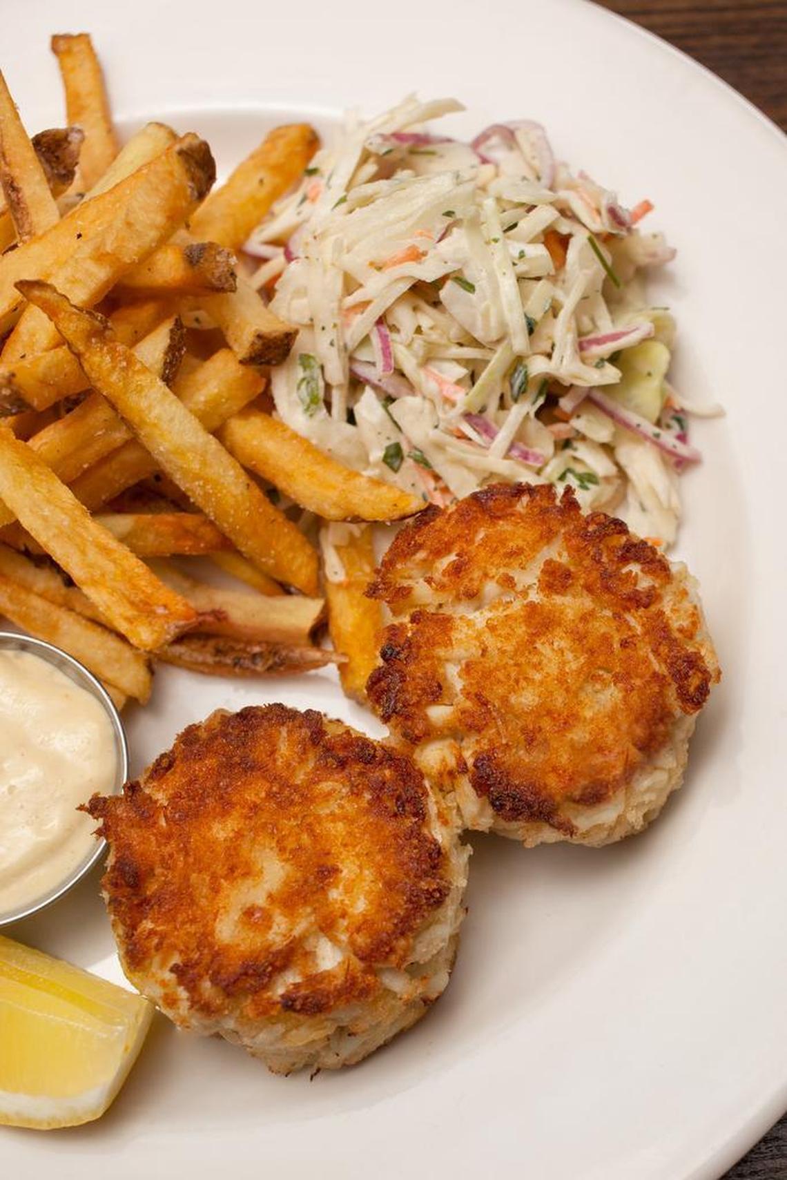 An overhead, close-up shot of a white plate featuring two golden-brown, pan-seared crab cakes. The crab cakes have a crispy, textured exterior and are served alongside a pile of thin-cut, skin-on french fries and a heap of creamy coleslaw with shredded cabbage and red onions. On the side, there is a small metal ramekin filled with tartar sauce and a fresh lemon wedge.
