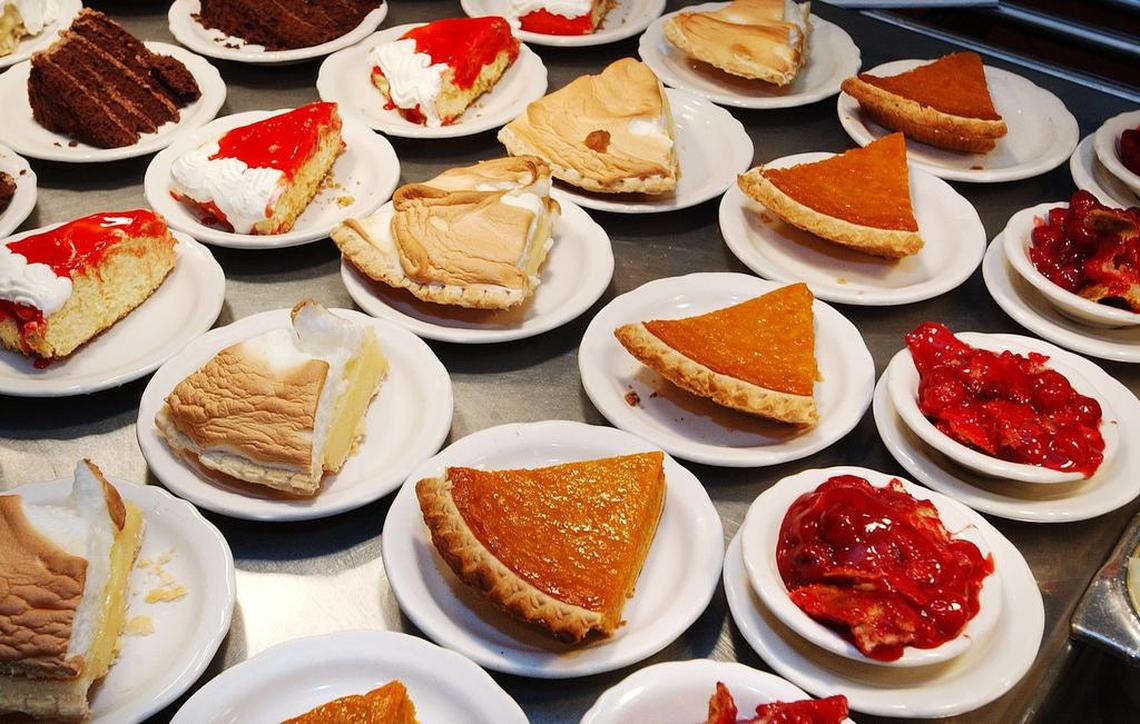 A high-angle, close-up shot of a counter displaying a dense cluster of various dessert slices on small white plates. The varieties include chocolate cake, lemon meringue pie, pumpkin pie, and cheesecake with red fruit topping.
