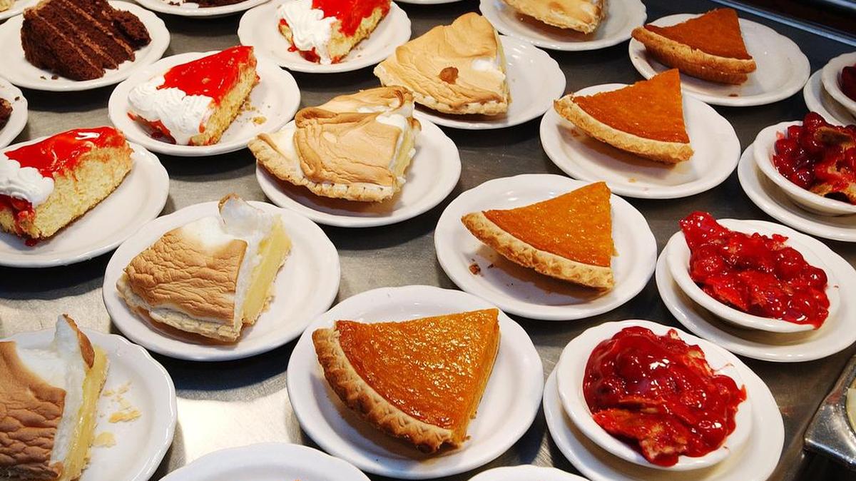 A high-angle, close-up shot of a counter displaying a dense cluster of various dessert slices on small white plates. The varieties include chocolate cake, lemon meringue pie, pumpkin pie, and cheesecake with red fruit topping.