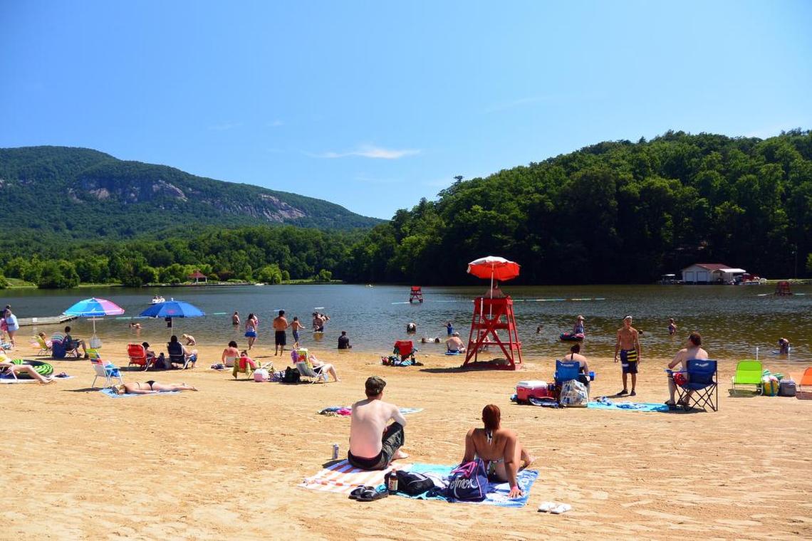 A sunny day at the beach on Lake Lure, viewed from the sand looking towards the water. In the foreground, a couple sits on a towel with their backs to the camera. The sandy beach is filled with other people relaxing, sunbathing under colorful umbrellas, and swimming in the calm lake. A red lifeguard stand sits at the water’s edge. The entire scene is surrounded by large, lush, tree-covered mountains under a clear blue sky.