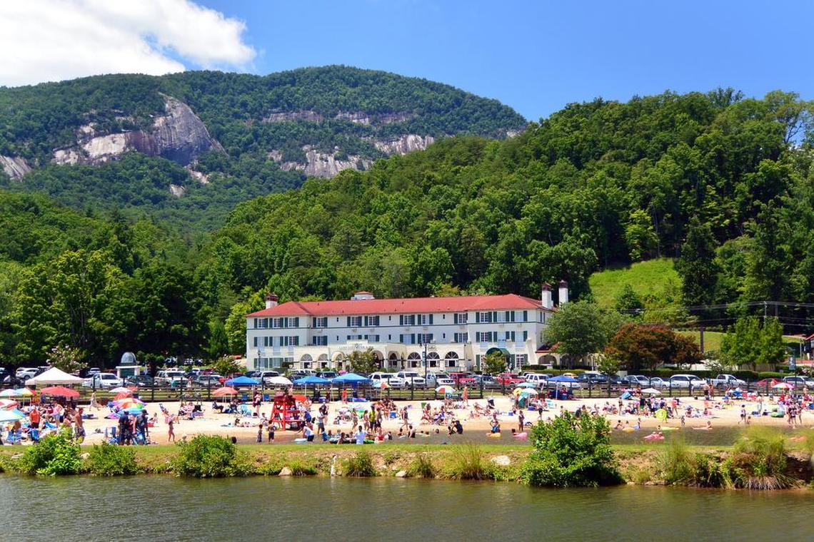 A wide, scenic shot of the Lake Lure Inn & Spa on a sunny day. In the foreground, a sandy beach is crowded with people sunbathing and swimming under colorful umbrellas. Behind them stands the large, historic white inn with a red roof, nestled at the base of a vast, tree-covered mountain with exposed rock cliffs under a blue sky with white clouds.
