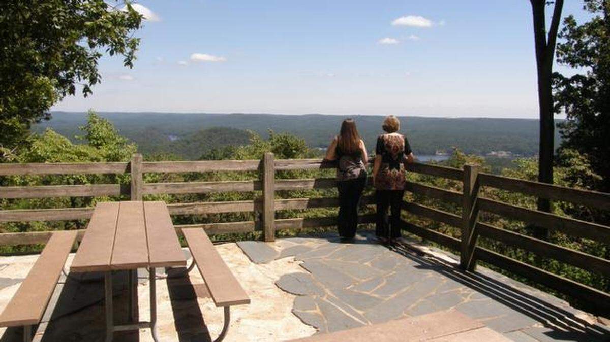 Two park visitors stand at a scenic overlook with a picnic table at Morrow Mountain State Park, looking out over a vast expanse of green, rolling hills and a distant lake under a blue sky.