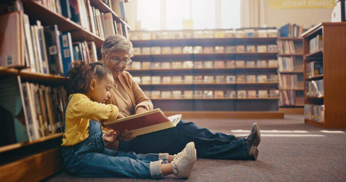 A grandmother with her grandchild sitting on floor of a library reading a book together. 
