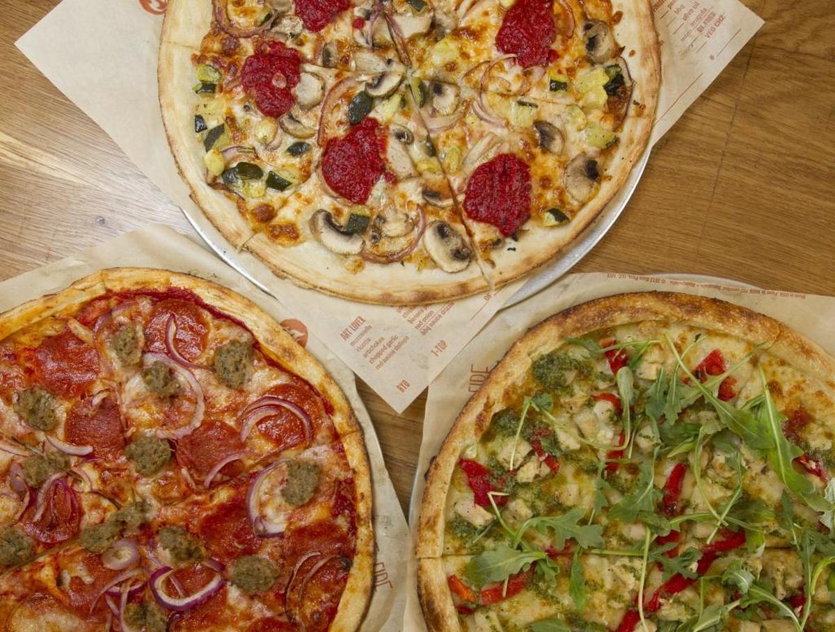 An overhead shot displays three different pizzas arranged on a light wooden table, each with a thin crust and various toppings. The pies include one with pepperoni, sausage, and red onion; one with pesto, chicken, red peppers, and arugula; and one with mushrooms, red onion, zucchini, and distinct dollops of thick red sauce.
