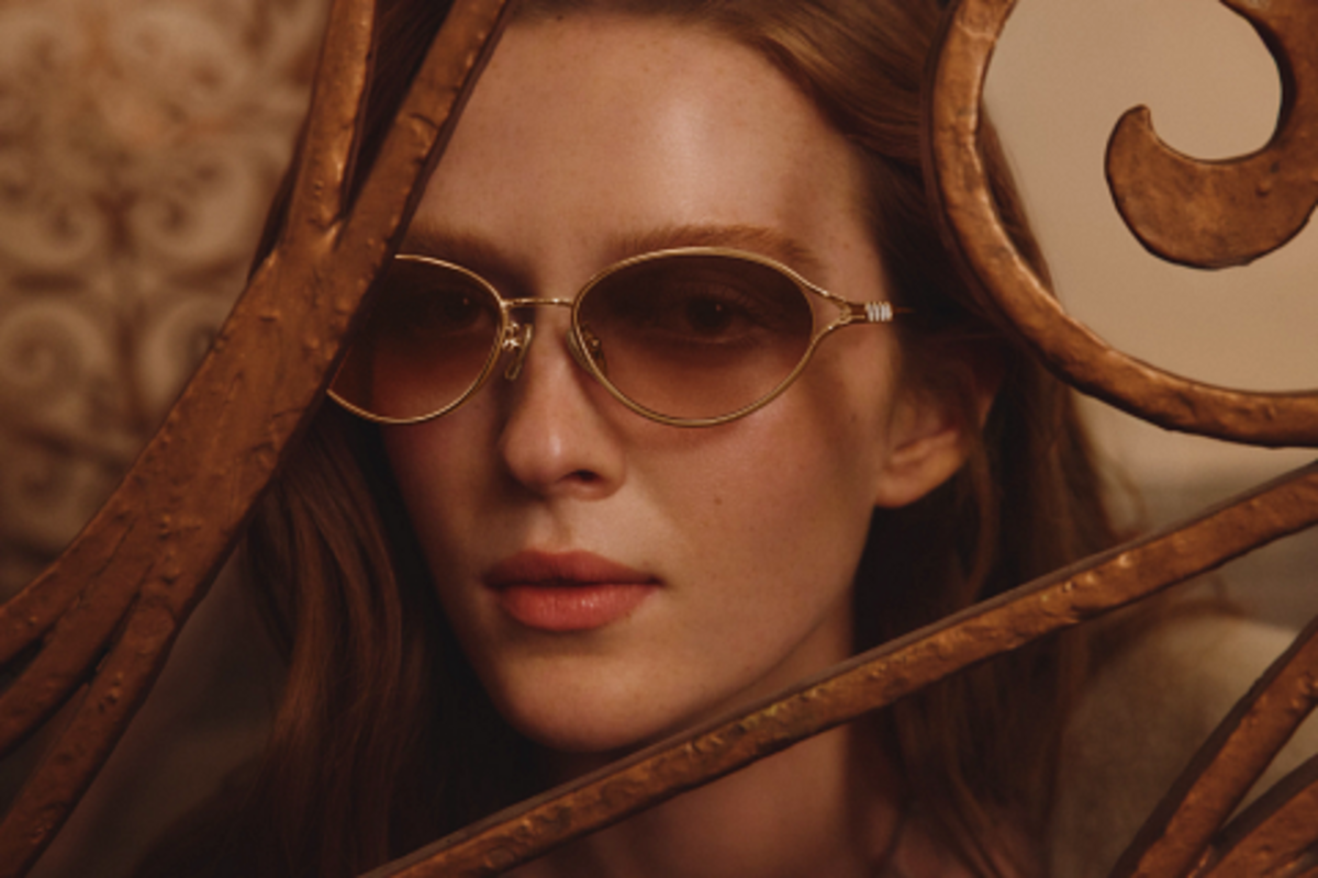 A close-up, softly lit portrait of a woman looking directly at the camera while wearing delicate, gold-rimmed oval sunglasses with brown-rose tinted lenses. She is partially framed by decorative, ornate golden metalwork or a gate, which adds a vintage or dramatic flair to the photo.
