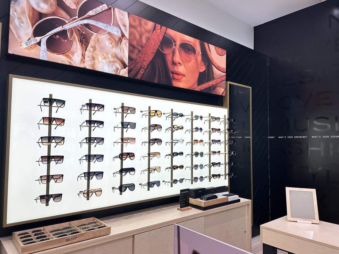 The second image captures a bright interior display wall of the QUAY store, showcasing dozens of sunglasses arranged neatly on lit vertical pegs. Below the large wall display are light wood cabinets holding more sunglasses trays and a small square mirror.