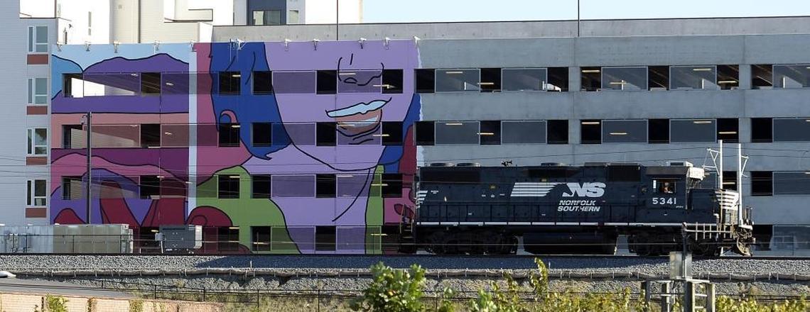 A locomotive passes by part of a mural that’s being painted on the side of the garage at the Novel NoDa apartment building on Tuesday, October 17, 2017.