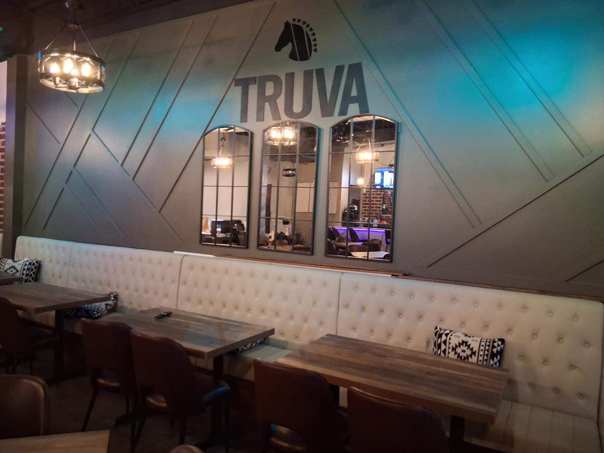 A restaurant interior featuring a long, tufted booth, several wooden tables, and dark leather chairs. Above the seating, the word TRUVA is displayed on a textured wall below a stylized horse head logo, flanked by three arched mirrors.