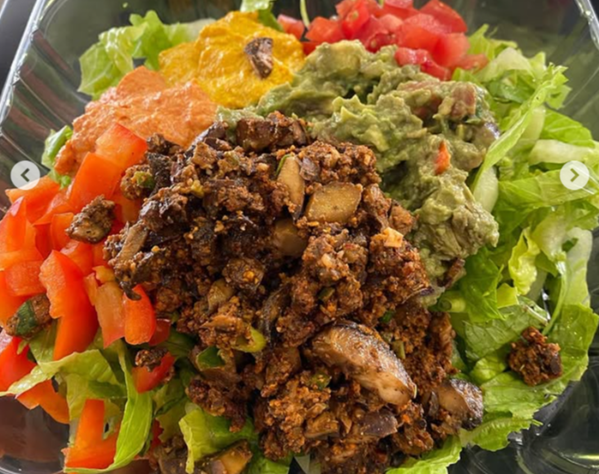 A close-up of a vibrant, colorful salad or bowl in a clear, square plastic to-go container. The base is chopped green lettuce, topped with a large mound of dark brown, savory plant-based “meat” crumble and sliced mushrooms. Surrounding the crumble are distinct toppings: diced red bell peppers on the left, a scoop of orange sauce or puree, a small scoop of yellow sauce or puree, a large scoop of chunky guacamole, and diced tomatoes on the top right.