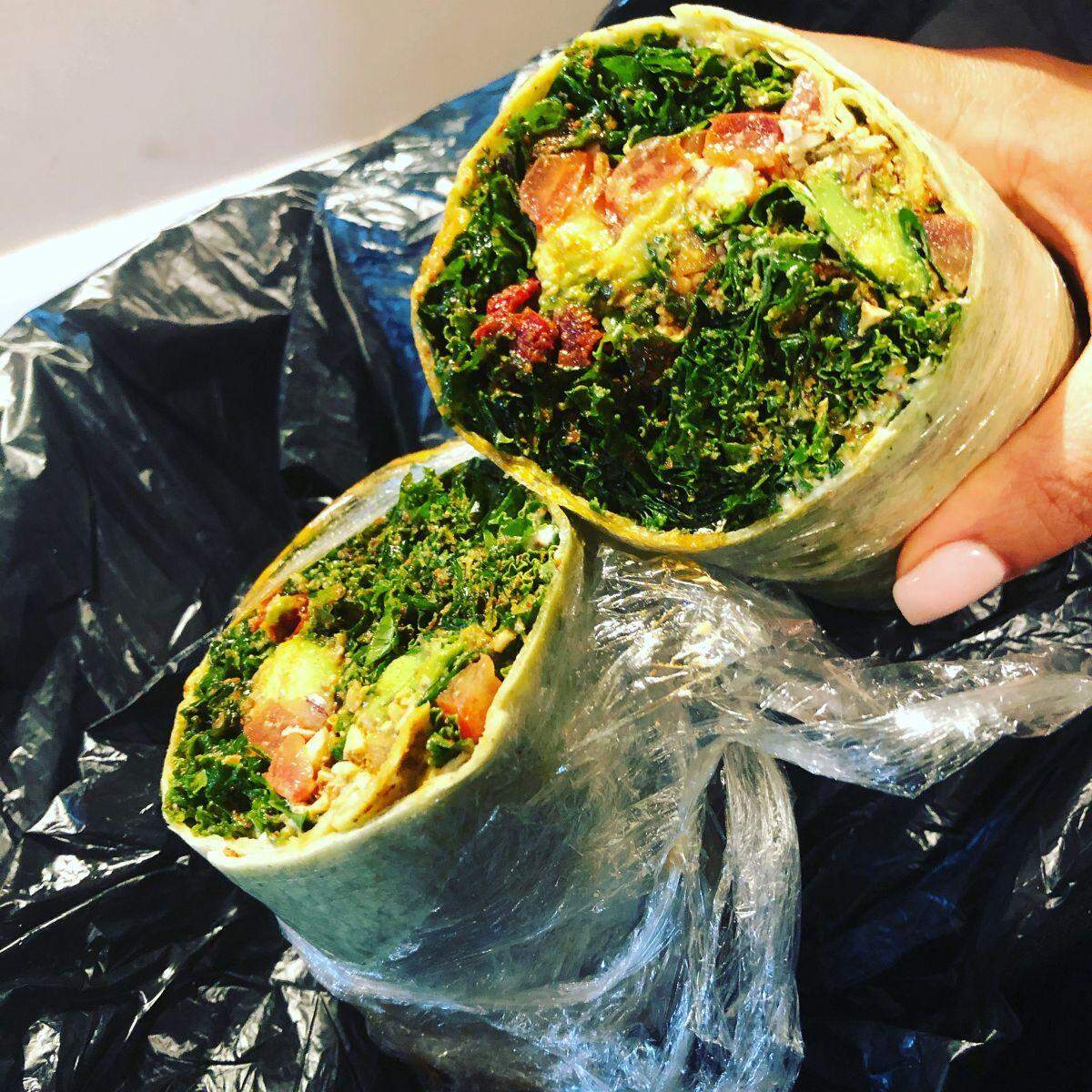 A hand holding a large, sliced-in-half green-hued wrap, wrapped tightly in clear plastic film. The cross-section shows a very dense filling dominated by finely chopped kale, sun-dried tomatoes, diced avocado, and a chunky, plant-based filling.