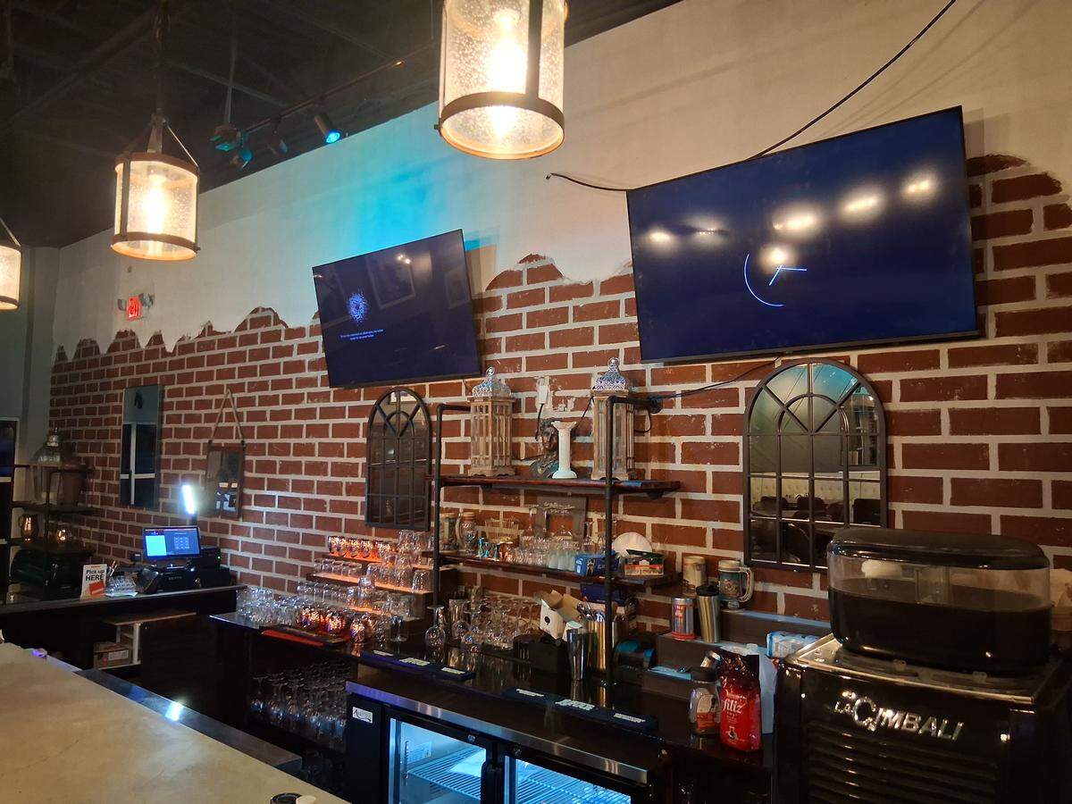 A close-up view of a restaurant bar area with a brick wall backdrop and two flat-screen TVs mounted above. The counter is lined with glassware, bar tools, and a large La Cimbali espresso machine on the right.