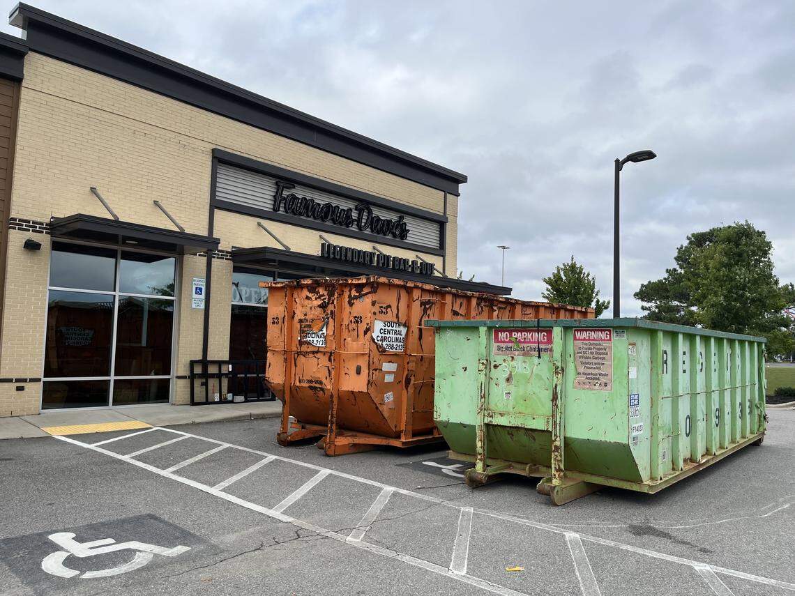 A wide-angle exterior photo showing a Famous Dave’s Legendary Pit Bar-B-Que restaurant, with two large, brightly colored construction dumpsters (one orange and one green) parked directly in front of the main entrance. The foreground includes a designated handicap parking space.