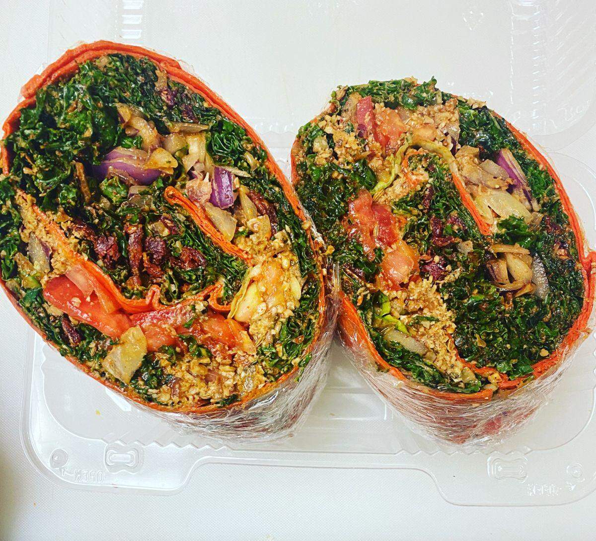 A large, brightly colored orange wrap, sliced in half, revealing a densely packed, vibrant filling. The filling consists primarily of chopped kale, sun-dried tomatoes, roasted vegetables like onions and peppers, and a textured, seed-and-nut-based crumble, all bound together with a light sauce.