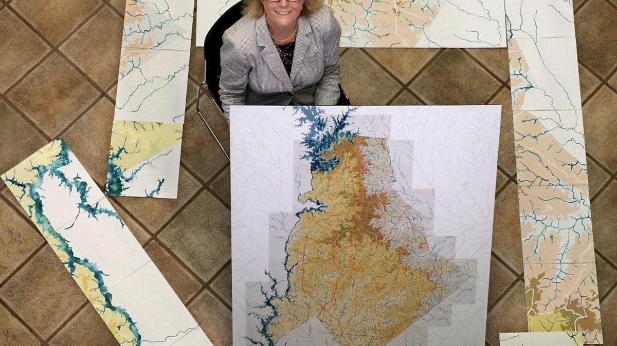 Charlotte Museum of History director Kay Peninger sits holding a copy of a replica map of the Waterways of Mecklenburg County, and she is surrounded by some of the sections of the larger project, a cut-paper map by Lauren Rosenthal which shows all of the county's streams and creeks, their systems and origins. OBSERVER FILE PHOTO