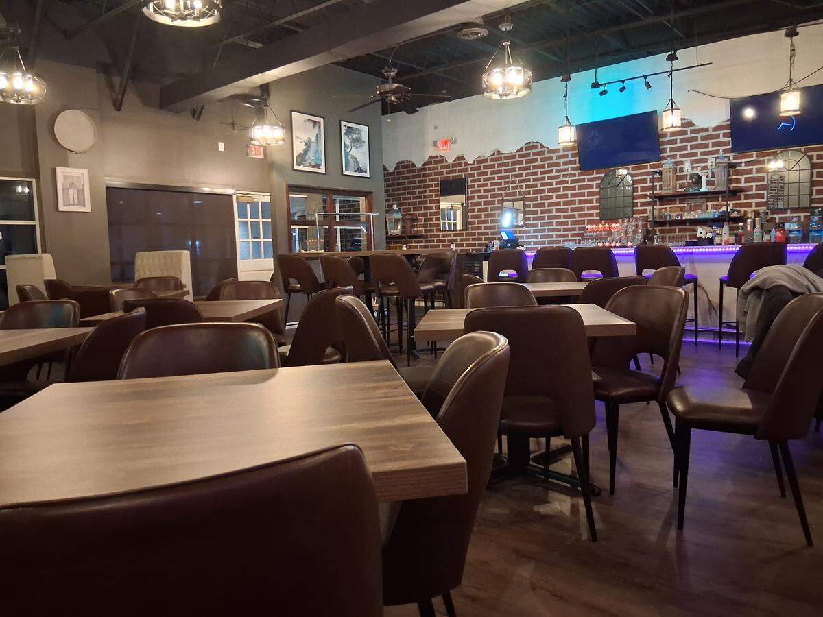 A view of a warmly lit dining room at night, featuring multiple square wooden tables and dark brown leather dining chairs. In the background, there is a bar area with a red brick wall and purple-lit countertop, lined with tall bar stools.