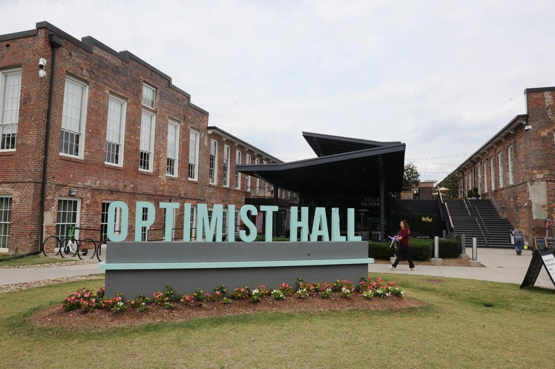 An eye-level, wide shot of the “OPTIMIST HALL” sign, which features large, light blue, sans-serif letters on a gray base, set on a grassy lawn with a flowerbed. In the background are the large, two-story, red brick former mill buildings that make up the hall, under an overcast sky.