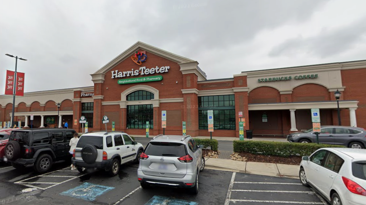 This wide, eye-level shot captures the exterior of a brick suburban shopping strip under an overcast sky. The main storefront features a large “Harris Teeter Neighborhood Food & Pharmacy” sign above an arched window entrance, while an attached “Starbucks Coffee” storefront sits directly to its right. In the foreground, several cars, including SUVs and sedans, are parked in an asphalt lot with visible blue accessible parking spaces.