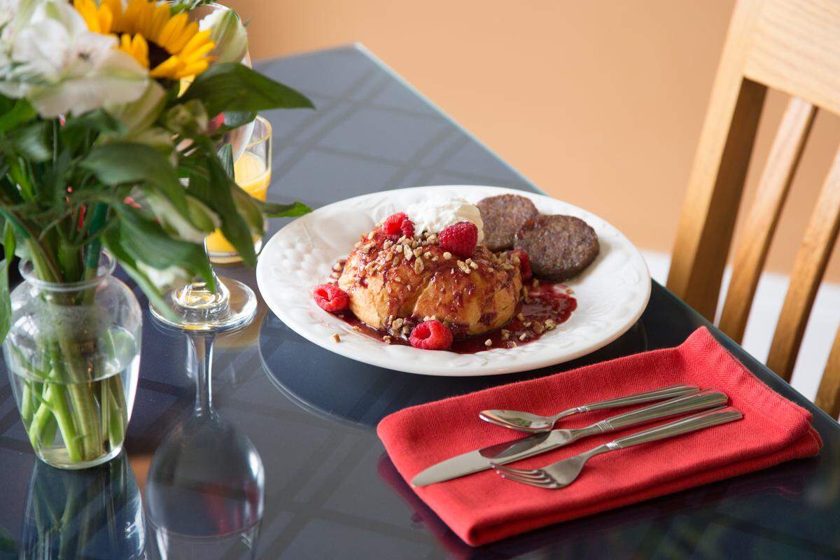 A plated breakfast of baked French toast with raspberries and pecans, topped with whipped cream and served with two sausage patties, is shown on a table. A glass of orange juice and a wine glass are in the background next to a vase of white and yellow flowers.