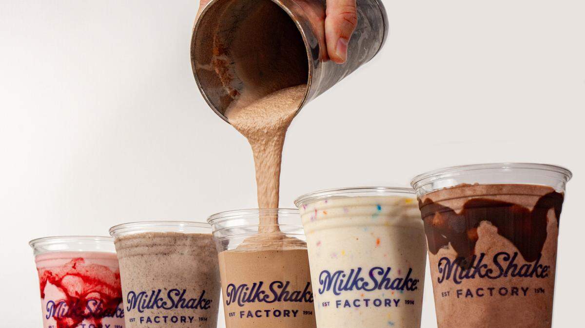 A hand holds a metal shaker and pours a chocolate milkshake into a clear plastic cup. Four other Milkshake Factory milkshakes in various flavors and toppings stand in a line on a surface.