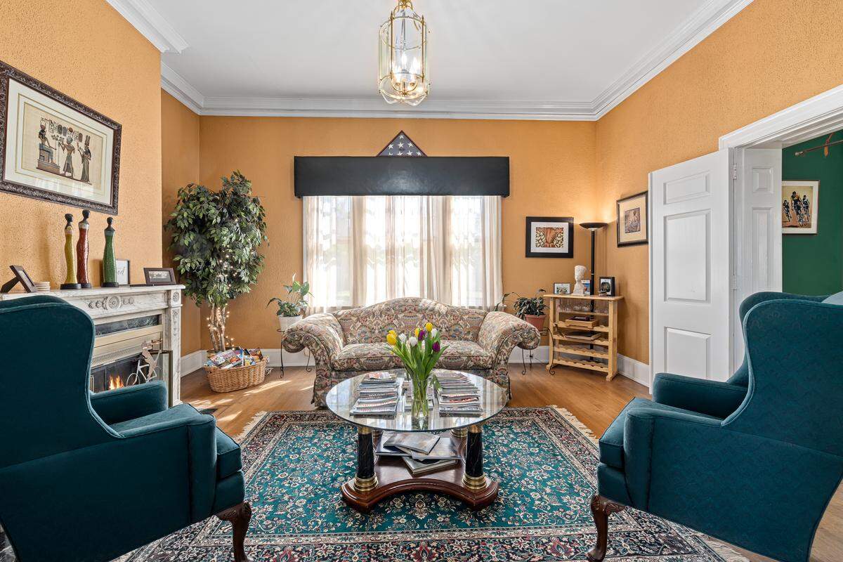 A traditional living room or library is shown with warm yellow walls and a fireplace. The room features a floral-patterned couch, two teal armchairs, and a round glass coffee table on a large Persian rug.