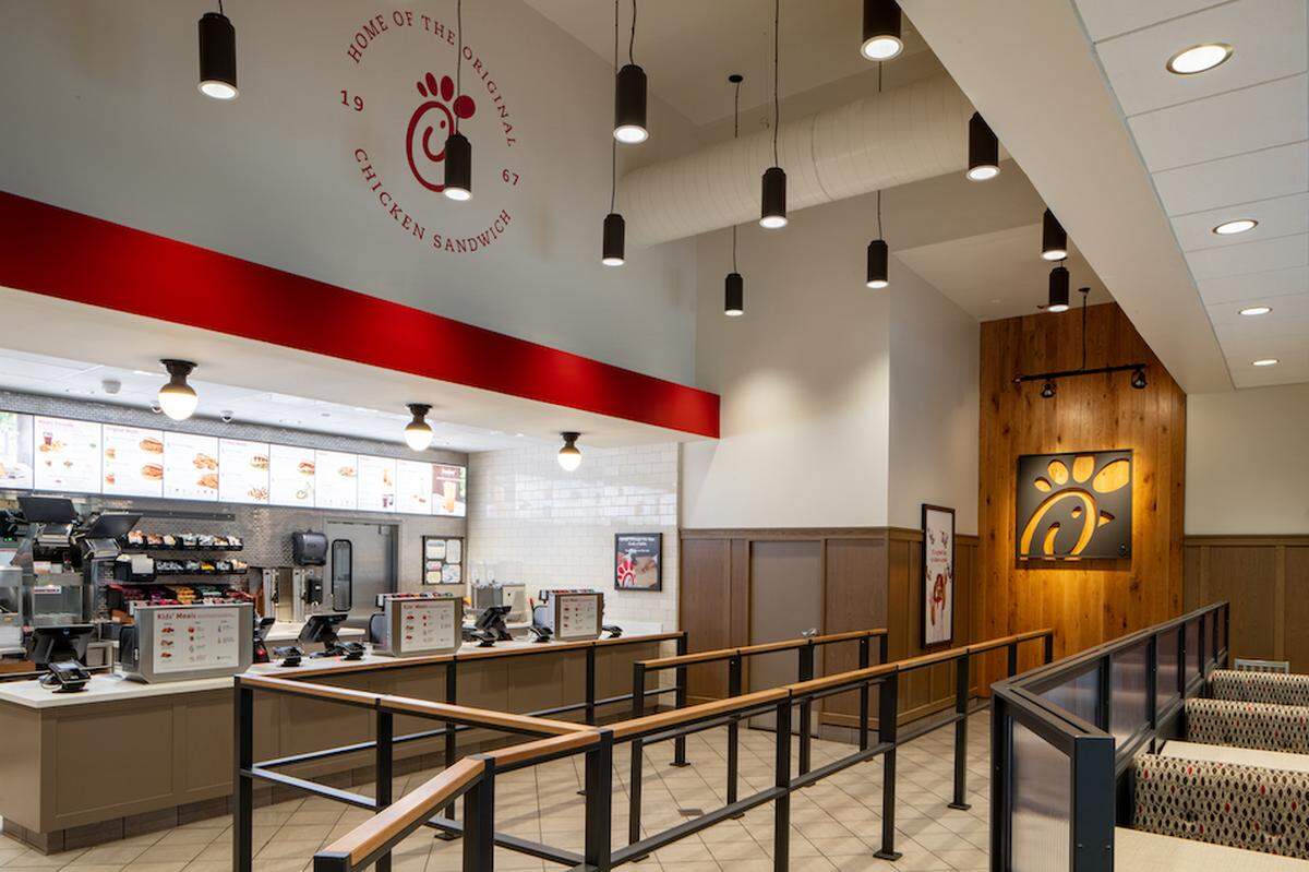 The interior of a Chick-fil-A restaurant shows the service counter with menus above it, and a roped queue leading towards the ordering area. The walls feature wood paneling and the Chick-fil-A logo, while modern pendant lights hang from the ceiling.