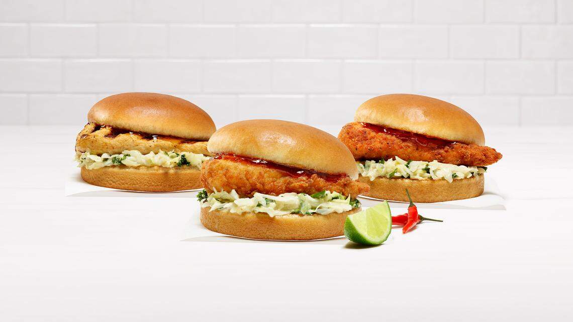 Three chicken sandwiches are arranged on a white surface, showcasing grilled, original, and spicy fillets topped with a dark sweet chili sauce and lime-infused slaw. A fresh lime wedge and small red chili peppers sit in the foreground against a clean, white subway tile background.
