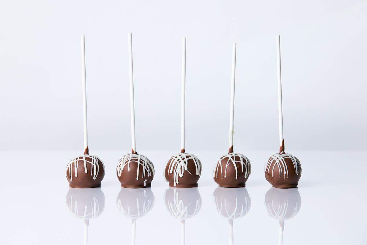 Five chocolate cake pops are arranged in a line on a white surface, with a white chocolate drizzle on each one. The cake pops are on white sticks, and their reflections are visible on the shiny surface below.
