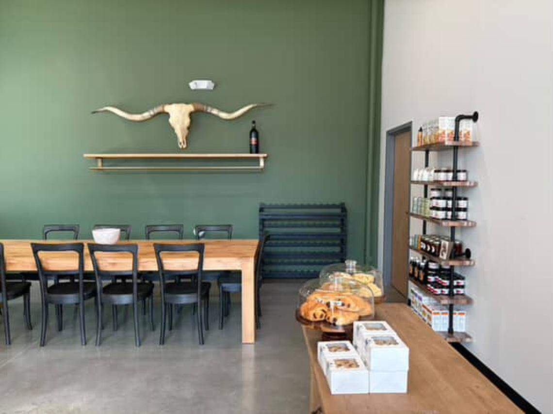 A modern cafe interior features a long communal wooden table with black chairs, and a small counter area with pastries in glass domes. A large set of longhorn cattle horns hangs on a green accent wall above a small floating shelf with a wine bottle.