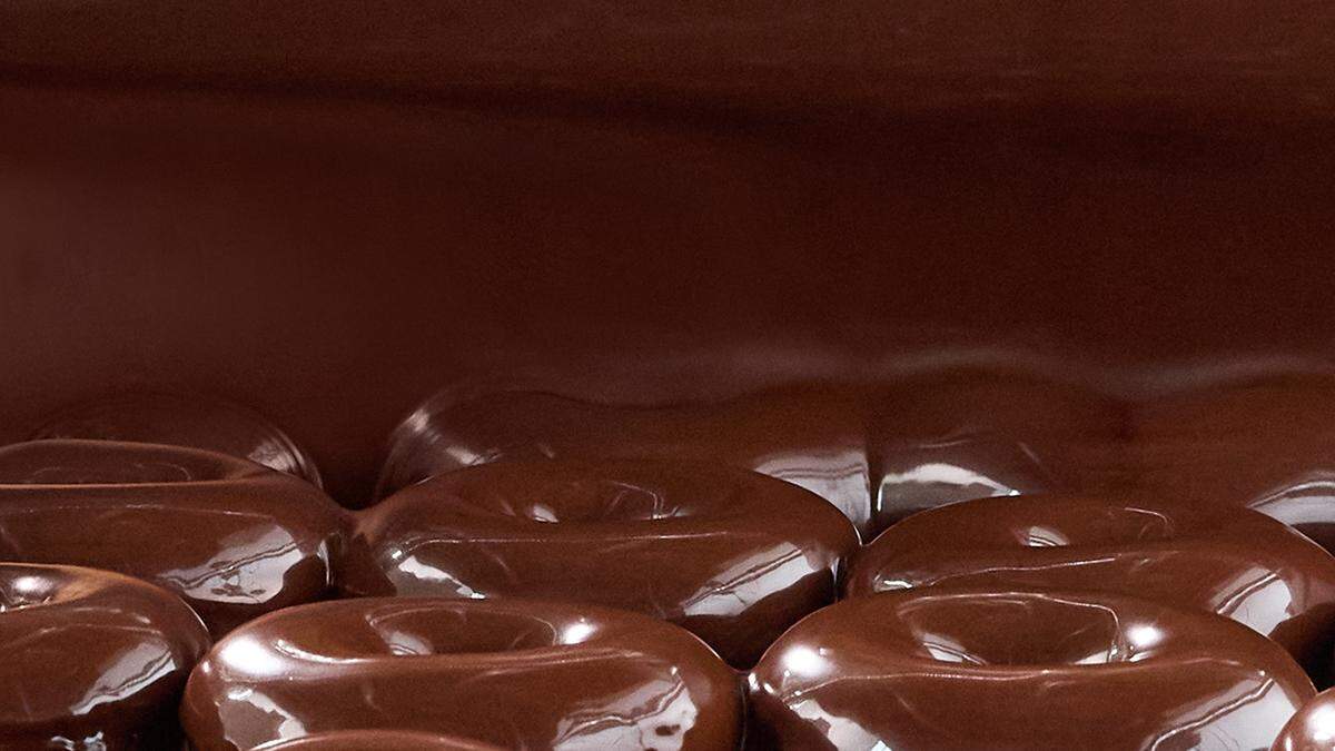 A close-up view shows a line of Krispy Kreme doughnuts being generously coated in a smooth, shiny chocolate glaze. The thick glaze flows over the tops and sides of the doughnuts, creating a rich, delectable appearance.