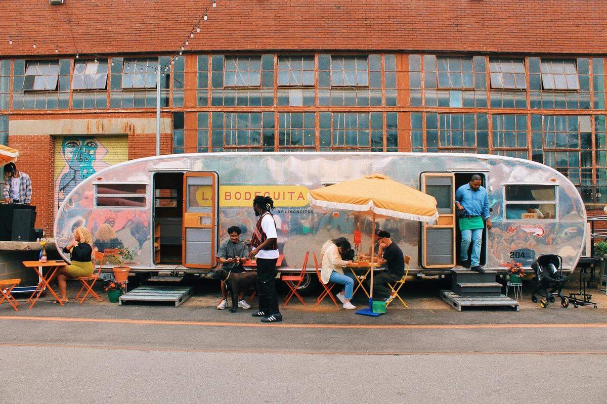 A vintage silver trailer with “La Bodeguita” written on it is parked outside a brick building with large windows. Several people are seated at small orange tables with chairs, and a yellow umbrella provides shade for a few of them.