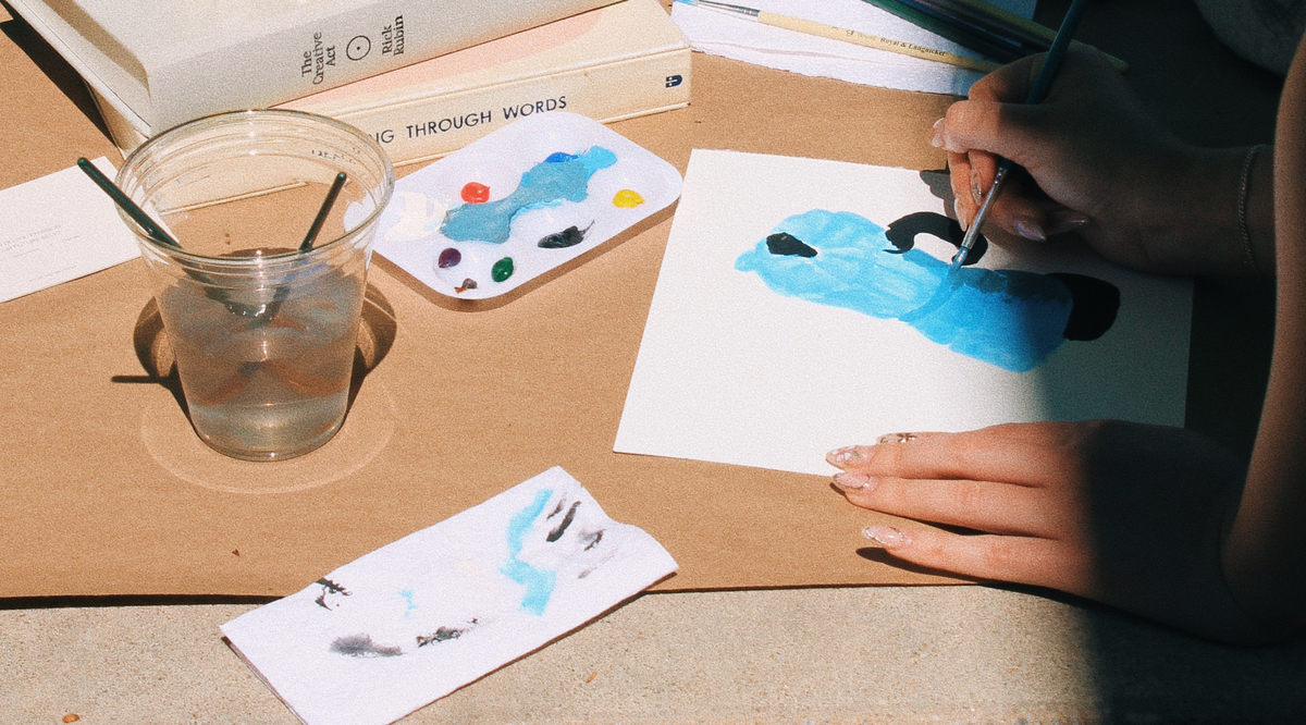 A person is painting a light blue figure with black details on a piece of white paper. Next to the painting is a plastic palette with various colors of paint, a plastic cup of water with paintbrushes inside, and a paper napkin with a smudge of blue paint.