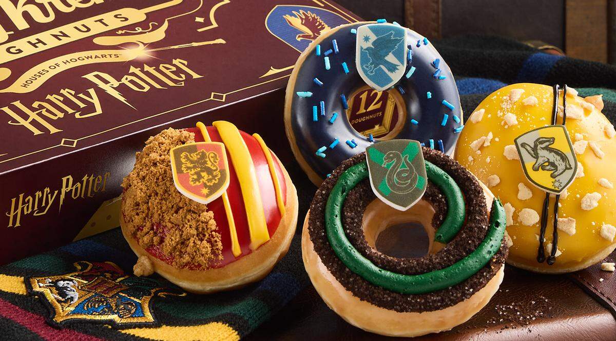 A box of Harry Potter-themed Krispy Kreme Doughnuts is displayed with four decorated doughnuts in front of a vintage suitcase. Each doughnut represents one of the four Hogwarts houses: Gryffindor, Ravenclaw, Hufflepuff, and Slytherin.