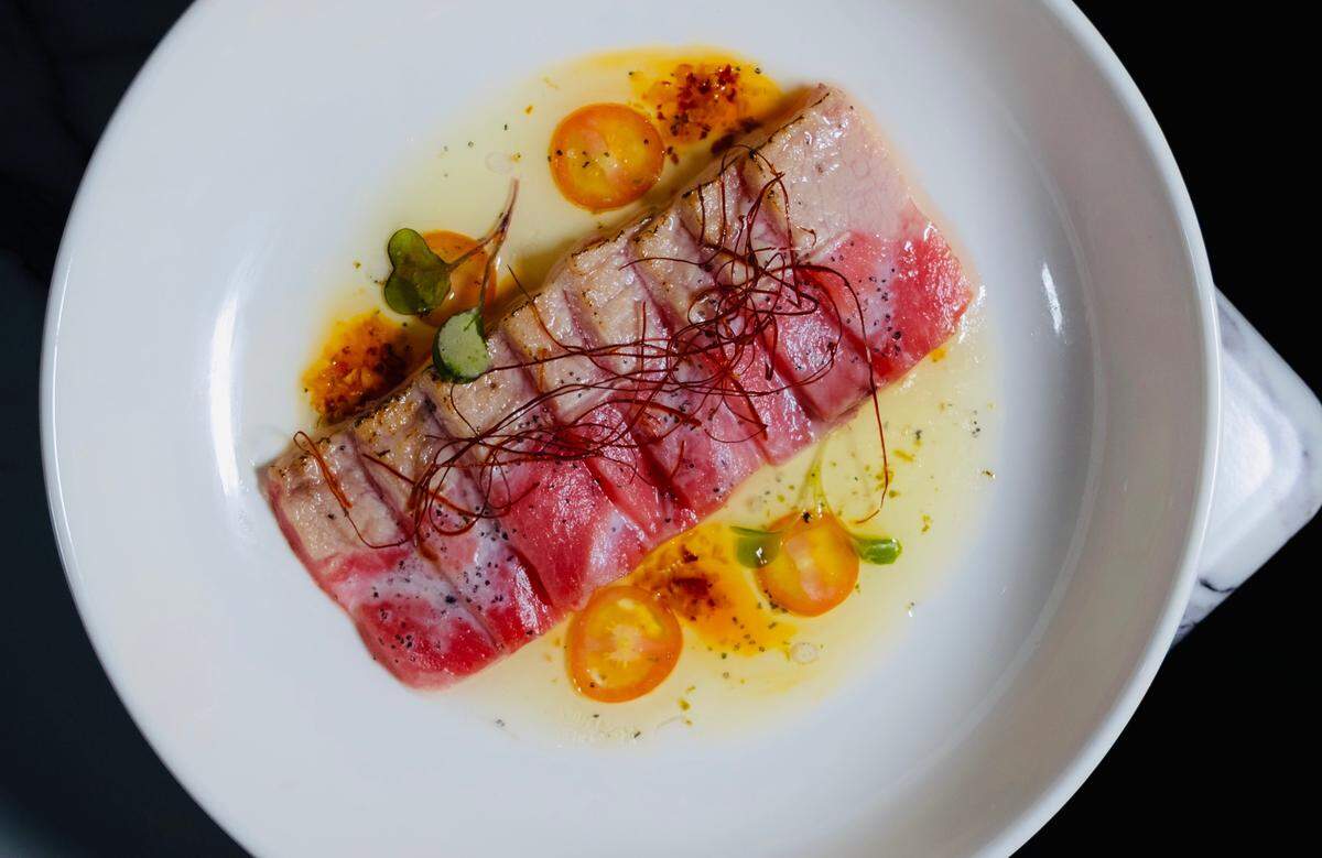 A top-down view of a rectangular piece of seared tuna on a white plate. It is garnished with a small amount of microgreens, thin red chili threads, and scattered halved cherry tomatoes, all resting in a pool of clear sauce with red oil drops.