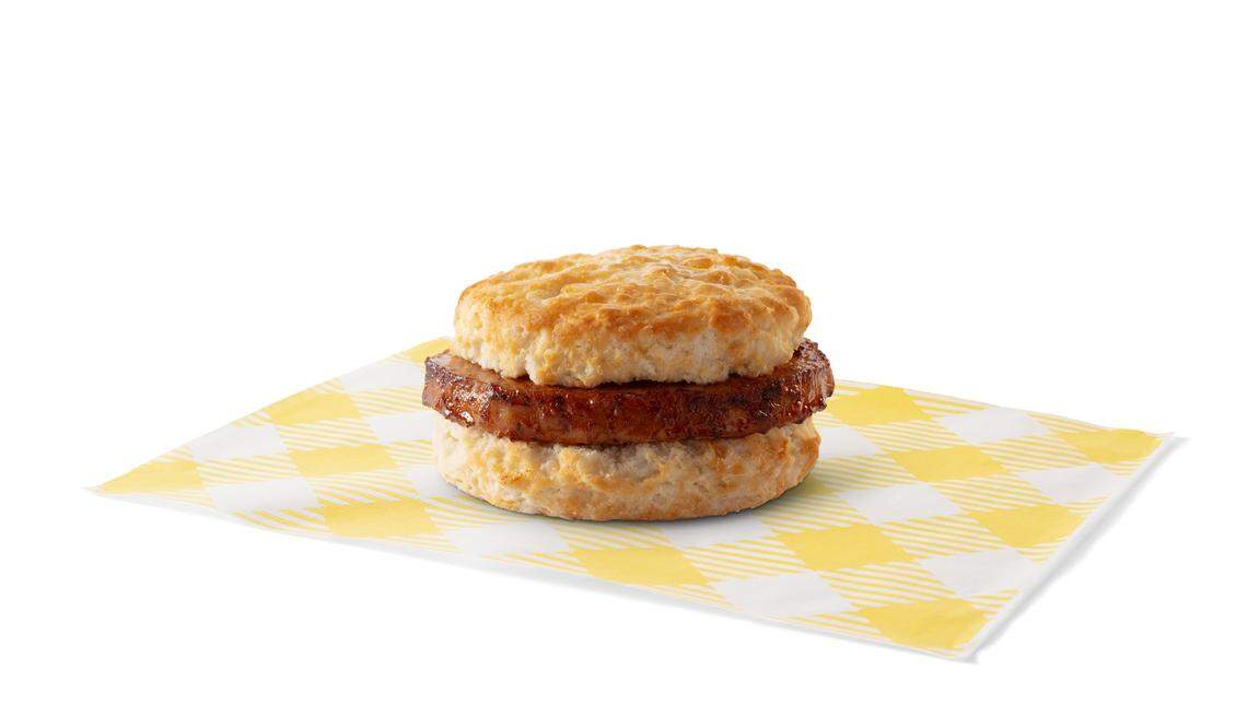 A golden-brown sausage patty is shown sandwiched between a flaky biscuit on a white and yellow checkered paper. The biscuit and sausage are centered on the paper with a white background.