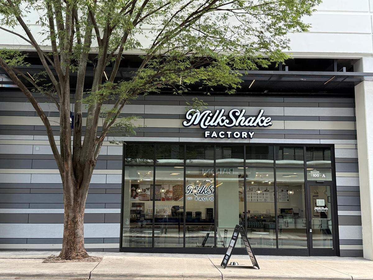 The front exterior of a Milkshake Factory store is visible in Charlotte, North Carolina. The storefront features large glass windows and a prominent lighted sign with the company’s logo.