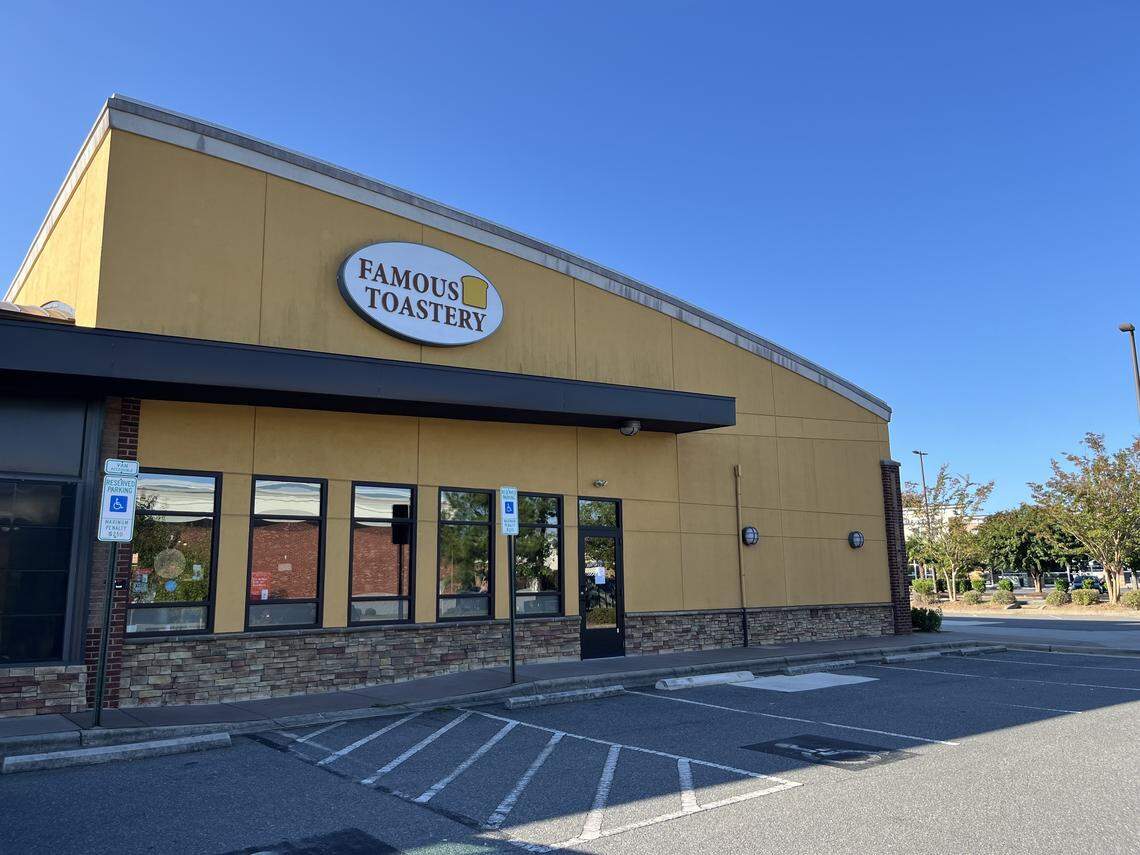 A wide, outdoor shot of the Famous Toastery restaurant, a single-story yellow building with a dark brown awning and a parking lot in the foreground. The building has a stone-patterned facade near the bottom, and the sky is clear and blue.