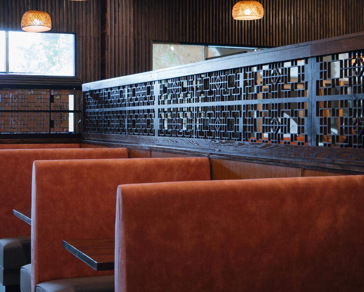 A series of empty restaurant booths with orange and gray cushions are separated by a dark wooden divider with a geometric pattern. Woven globe-shaped light fixtures hang from the ceiling.