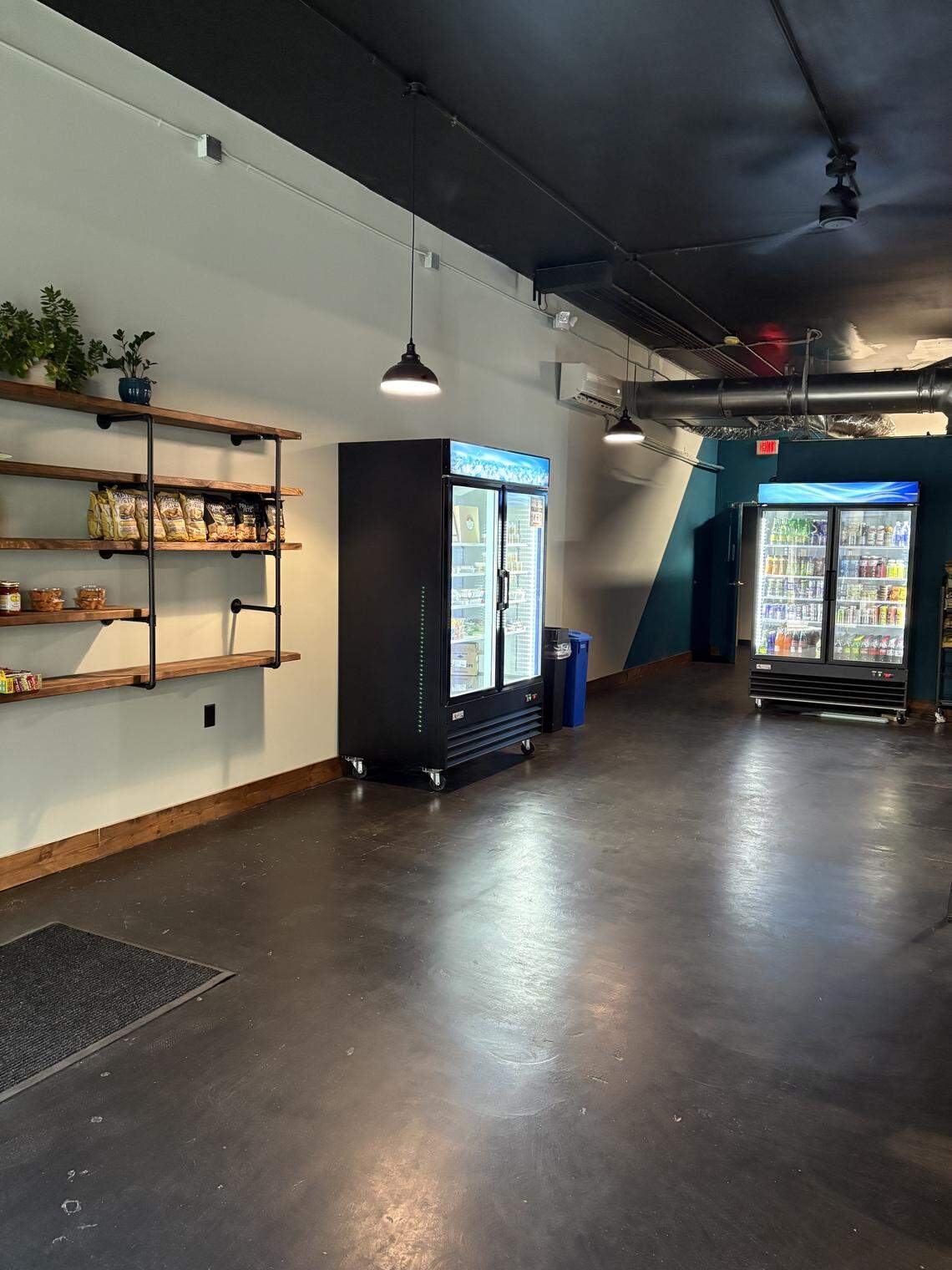 A photo shows a self-service beverage and snack area in a small eatery. There are two upright glass-front refrigerators filled with drinks, and a long shelf on the wall to the left is stocked with bags of chips and other snacks.